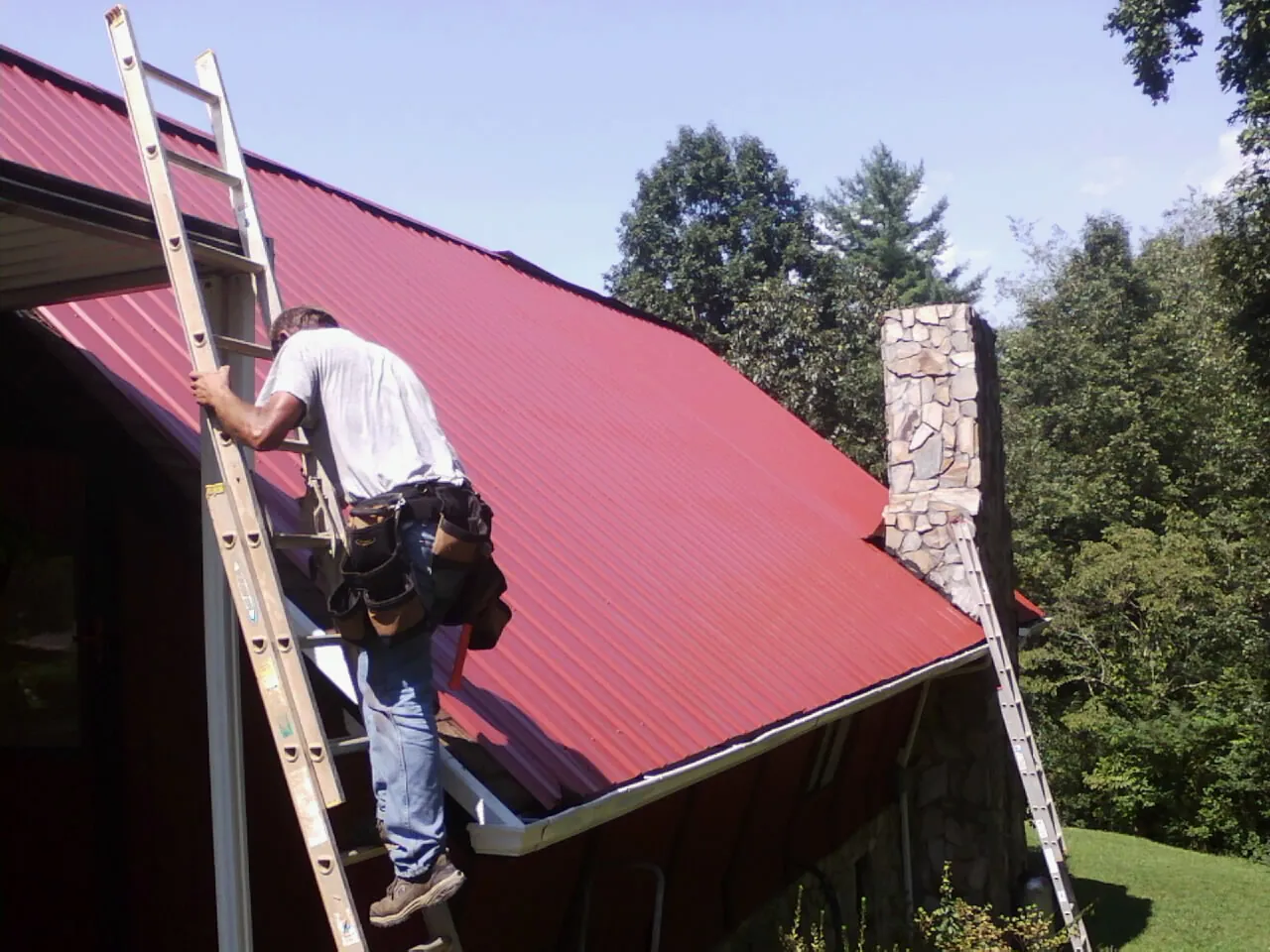 Roofer on a ladder beside a red metal roof and stone chimney — Soffit & Fascia Repair by Roofing Brothers in Polo